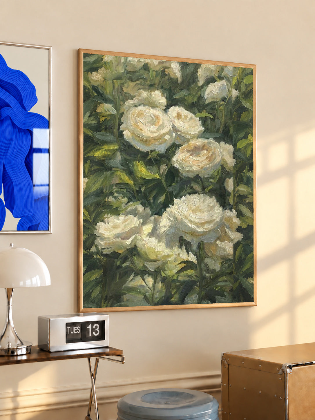 Cream white roses among dense green garden foliage in a painterly composition in an oak frame above a console beside a blue print and calendar