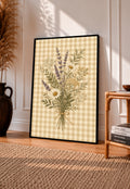 Tied bouquet of daisies lavender toned sprigs and white wildflowers on beige gingham in a black frame leaning on a sunlit wooden floor beside books and a rattan seat