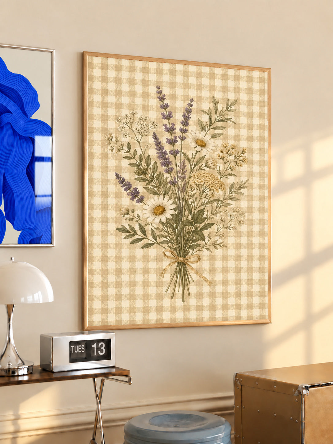 Tied bouquet of daisies lavender toned sprigs and white wildflowers on beige gingham in an oak frame above a console beside a blue print and calendar