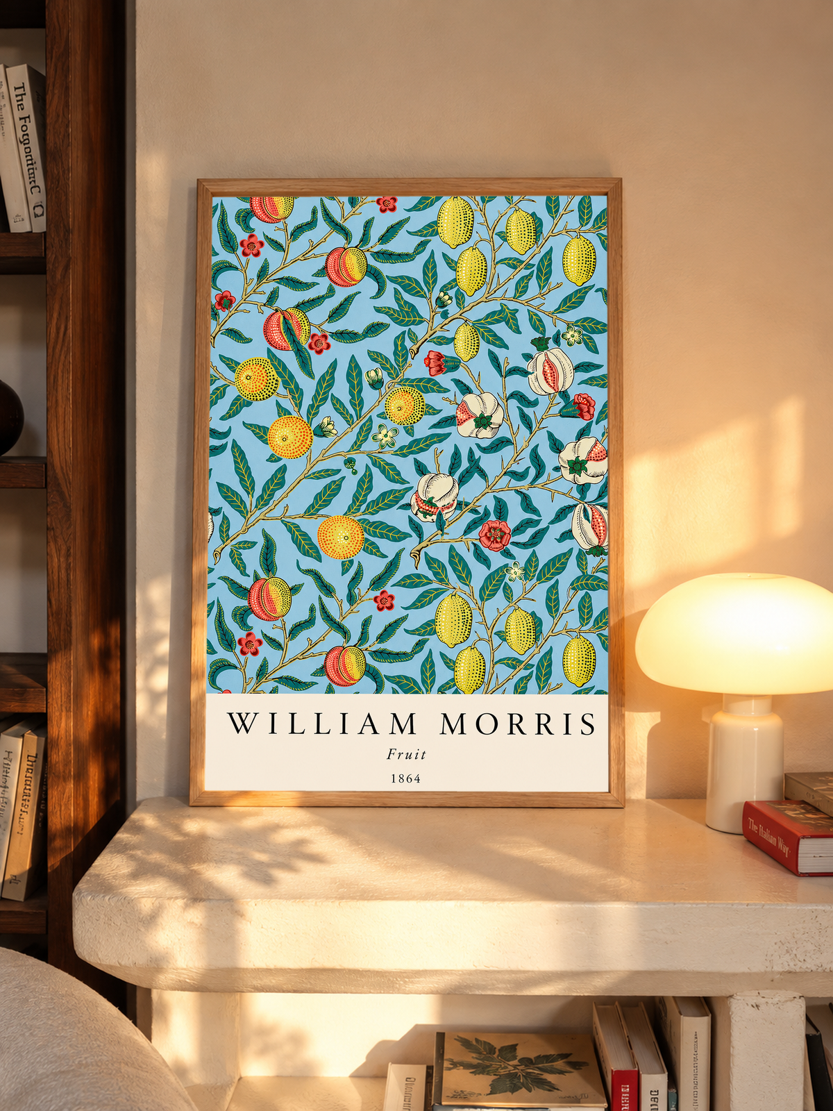 William Morris Fruit botanical pattern with yellow fruit and green leaves on pale blue in an oak frame on a sunlit console beside books and a cream lamp