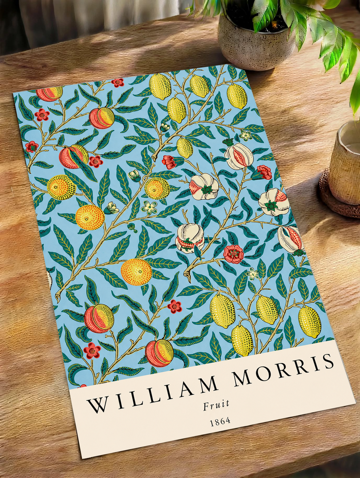 William Morris Fruit botanical pattern with yellow fruit and green leaves on pale blue as an unframed print on a wooden table beside a plant
