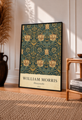 William Morris Honeysuckle floral pattern with gold and pink blooms on dark teal in a black frame leaning on a pale panelled wall beside a rattan chair