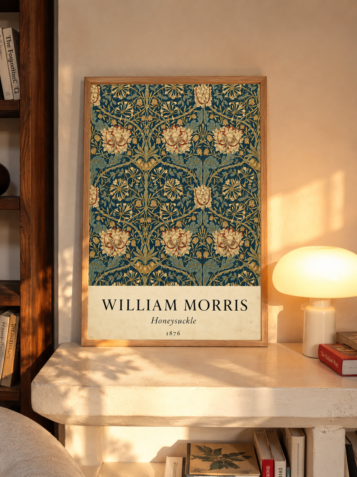 William Morris Honeysuckle floral pattern with gold and pink blooms on dark teal in an oak frame on a sunlit console beside books and a cream lamp