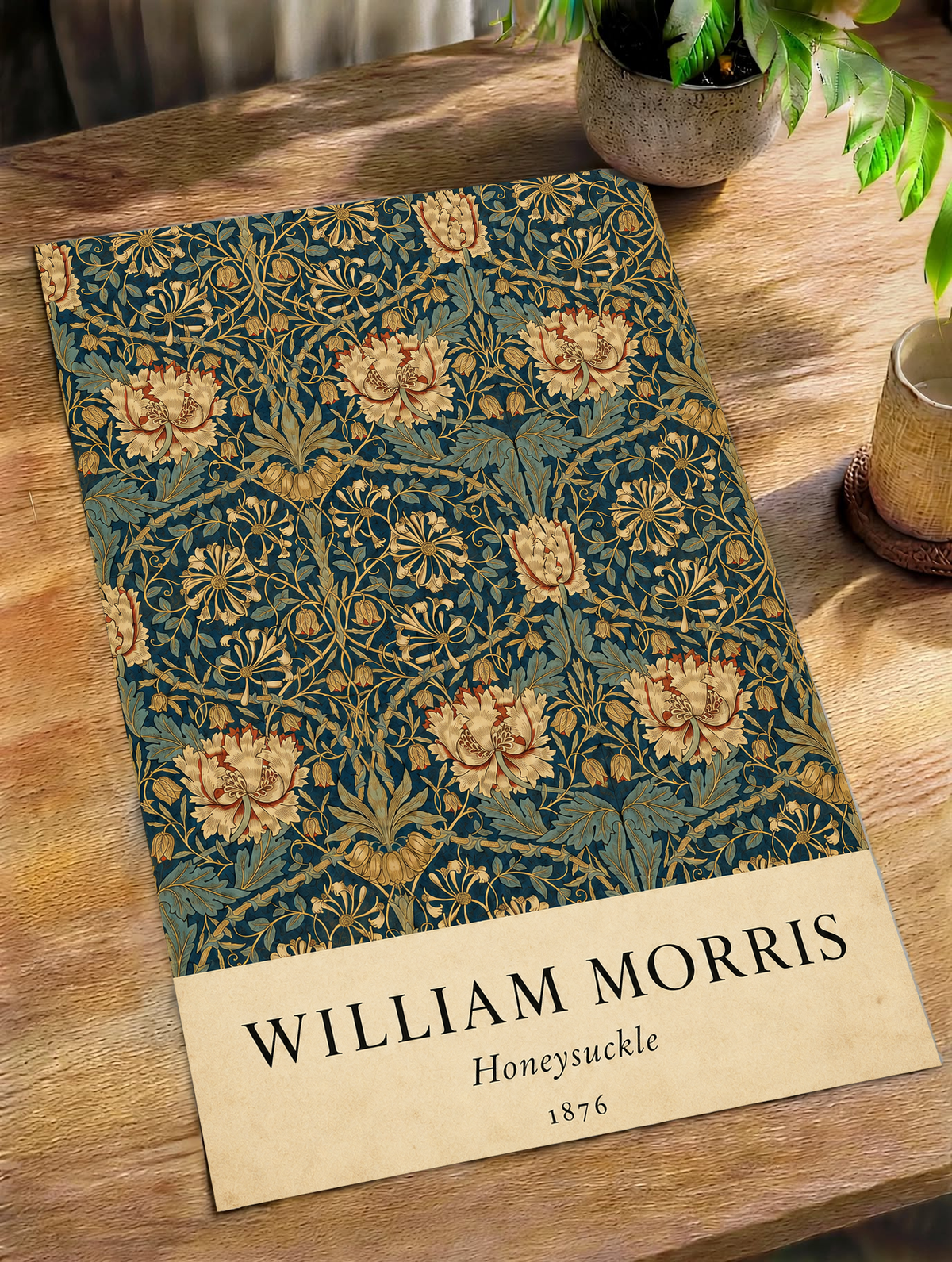 William Morris Honeysuckle floral pattern with gold and pink blooms on dark teal as an unframed print on a wooden table beside a plant