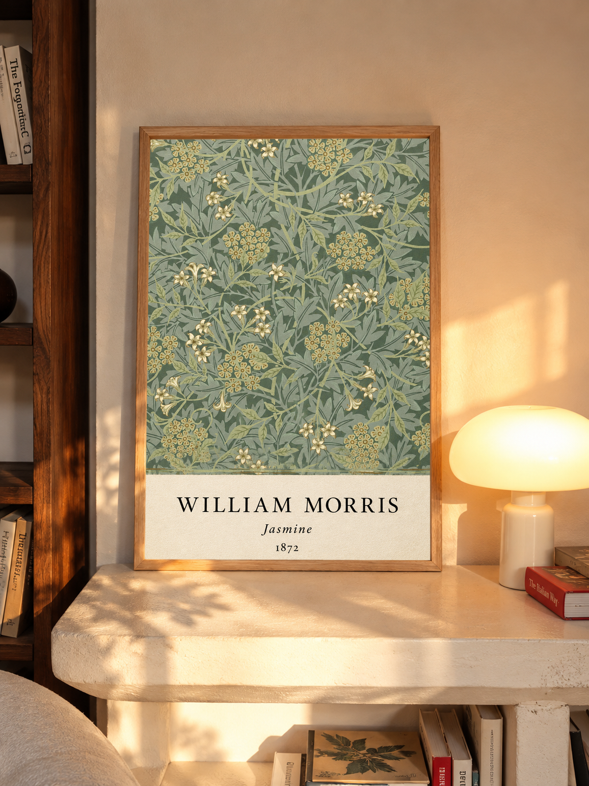 William Morris Jasmine sage and olive foliage with pale yellow flowers in an oak frame on a sunlit console beside books and a cream lamp