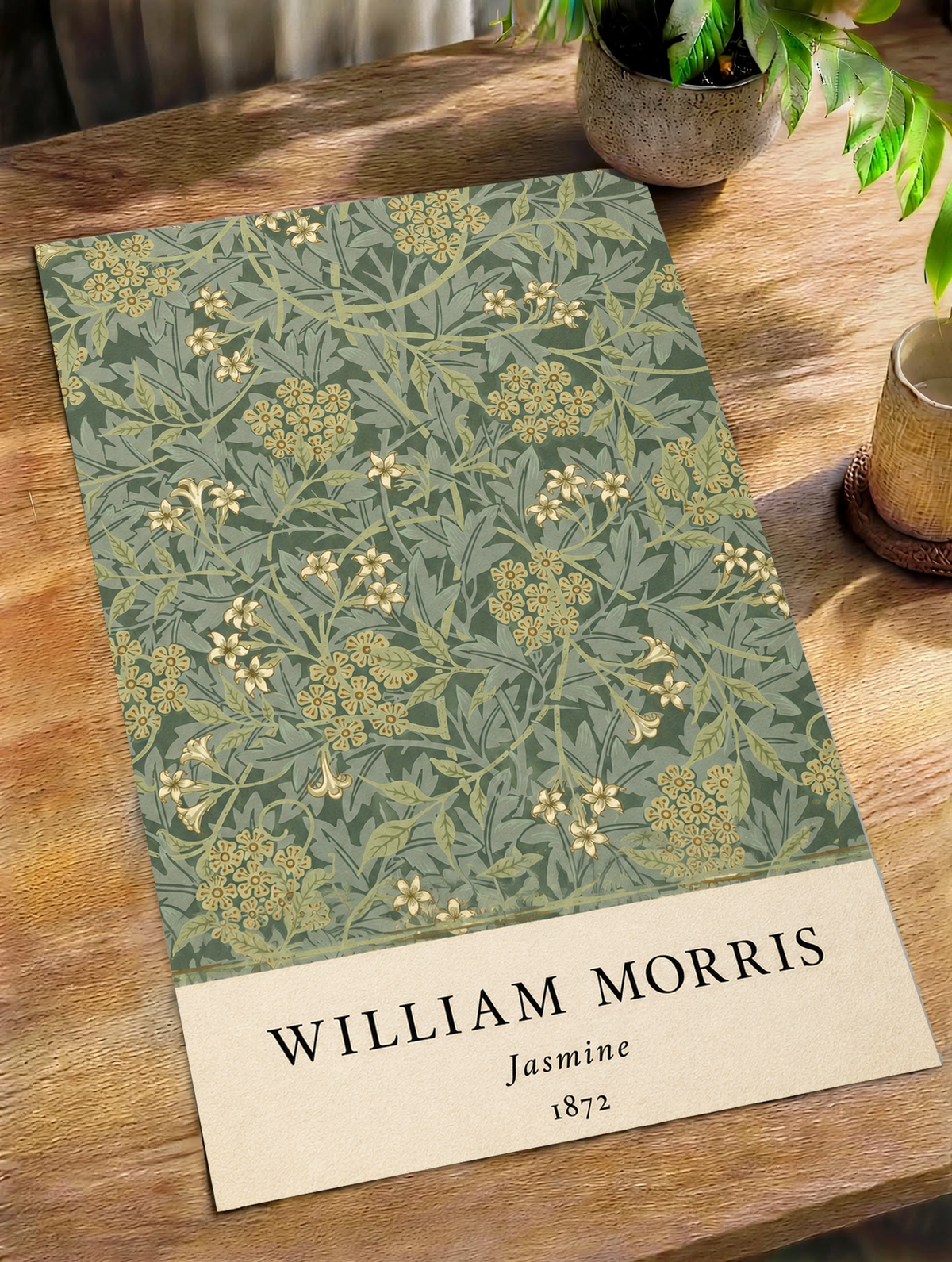 William Morris Jasmine sage and olive foliage with pale yellow flowers as an unframed print on a wooden table beside a plant