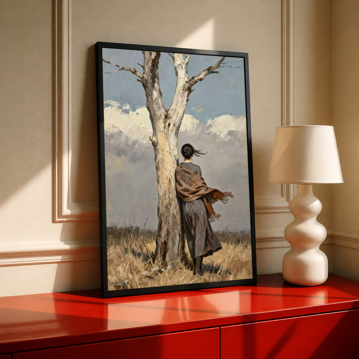 A woman in a long dark dress and brown shawl beside a pale tree in a windswept field in a black frame on a red console beside a sculptural cream lamp