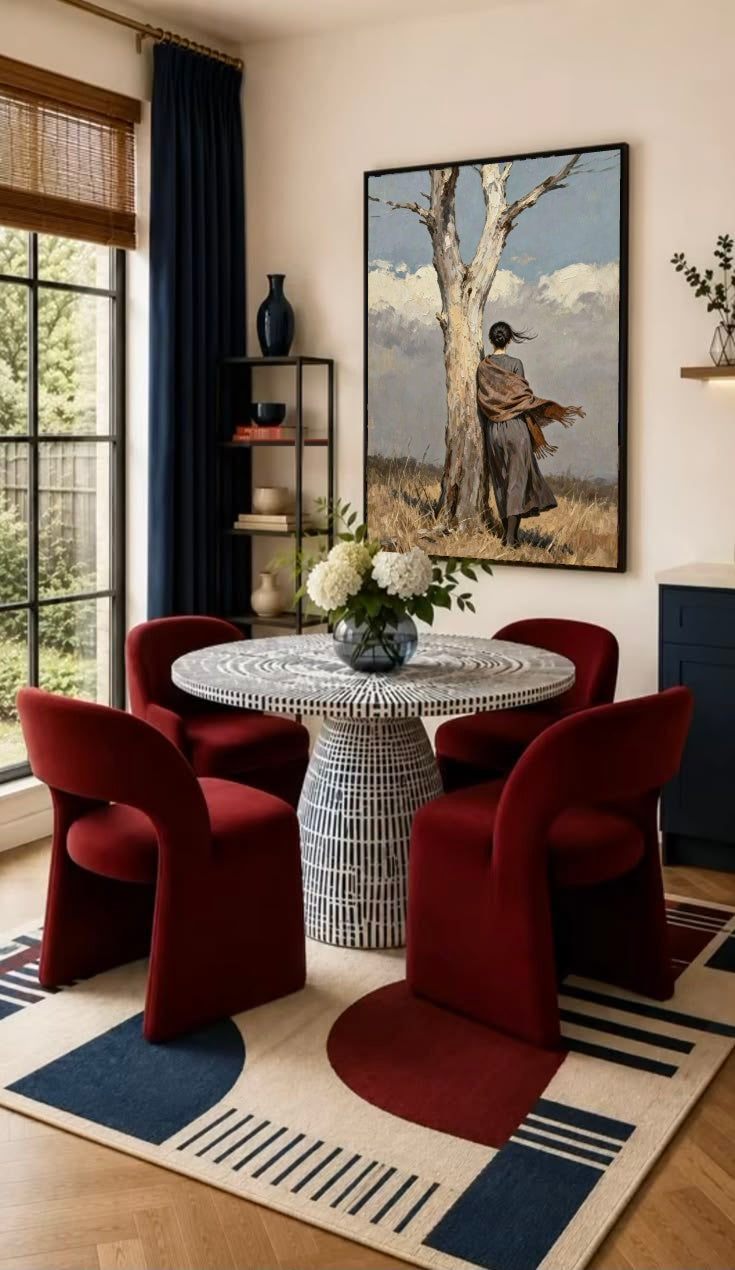 A woman in a long dark dress and brown shawl beside a pale tree in a windswept field in a black frame on the wall of a dining nook with red chairs
