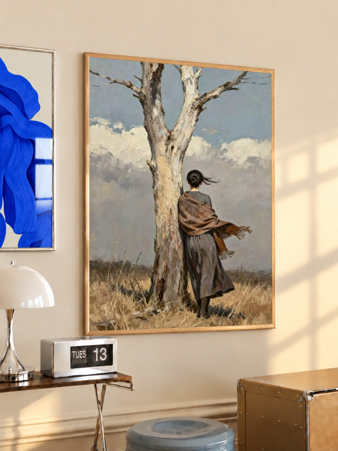 A woman in a long dark dress and brown shawl beside a pale tree in a windswept field in a light wood frame on a sunlit office wall beside a desk