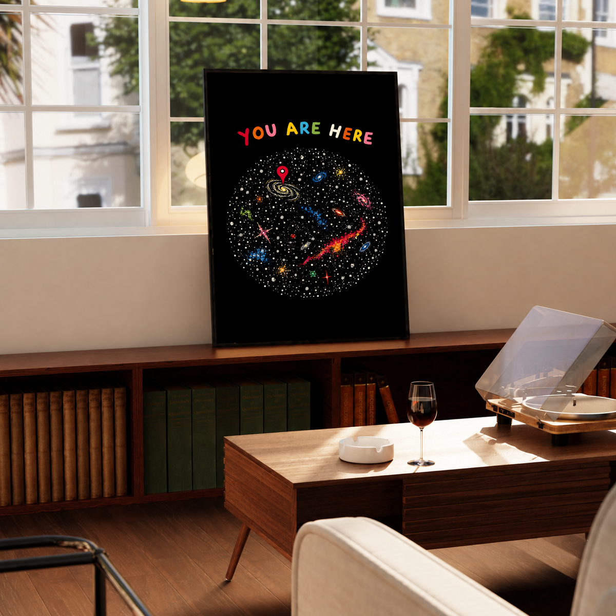 You Are Here multicolored lettering above a hand-drawn galaxy field with stars and a red location pin, styled in a black frame above a wood console with books and a record player