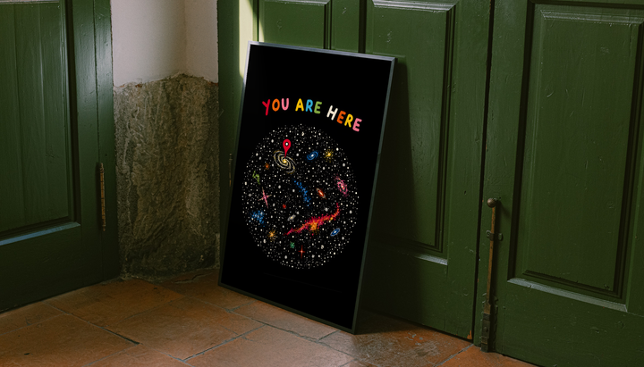 You Are Here multicolored lettering above a hand-drawn galaxy field with stars and a red location pin, styled in a black frame against dark green paneled doors above orange floor tiles