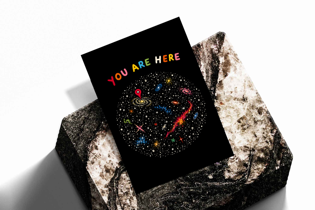 You Are Here multicolored lettering above a hand-drawn galaxy field with stars and a red location pin, shown as an unframed print tilted on a dark stone display against a light background