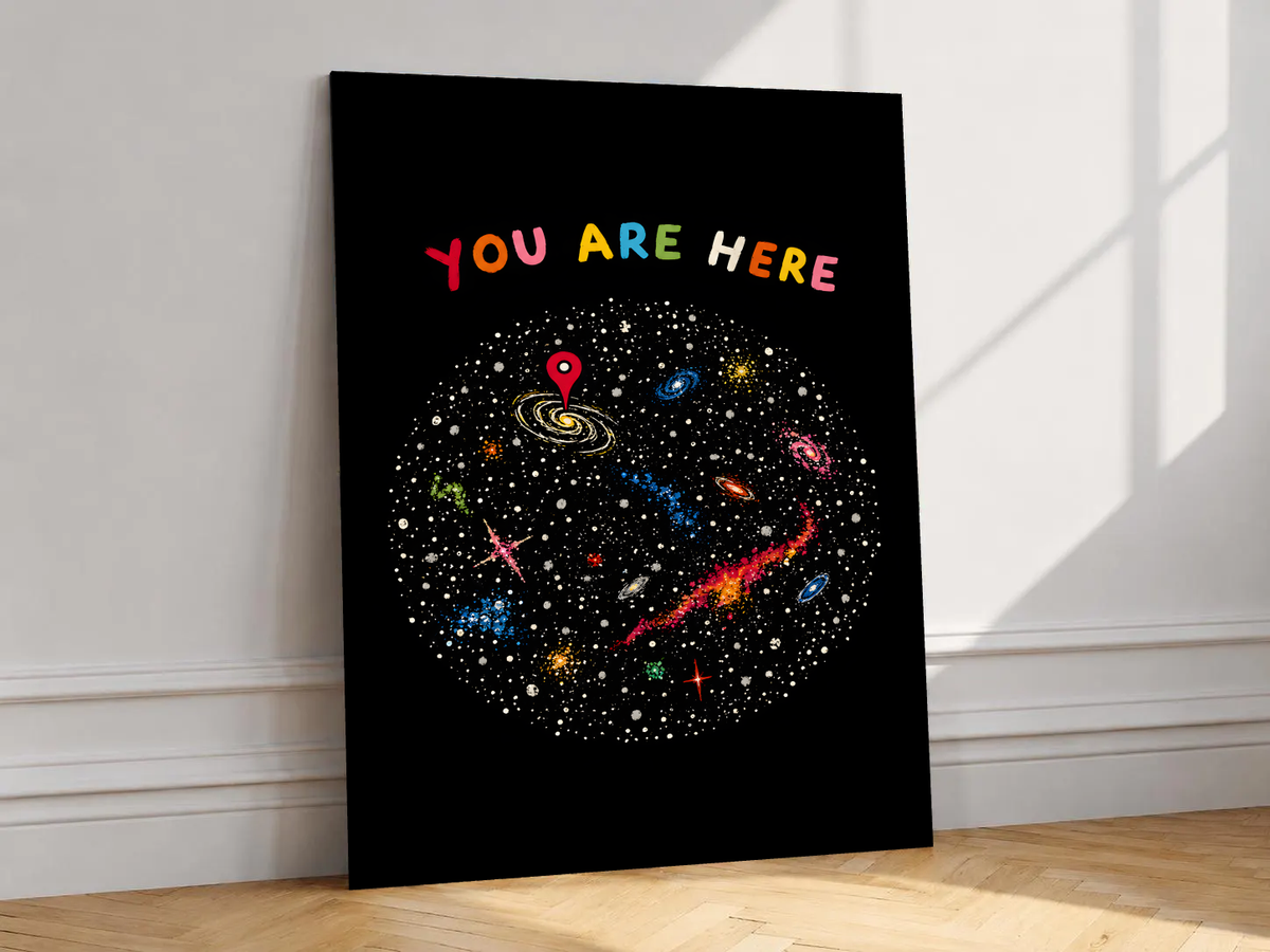 You Are Here multicolored lettering above a hand-drawn galaxy field with stars and a red location pin, shown unframed on a white wall above parquet flooring with a diagonal sun shadow
