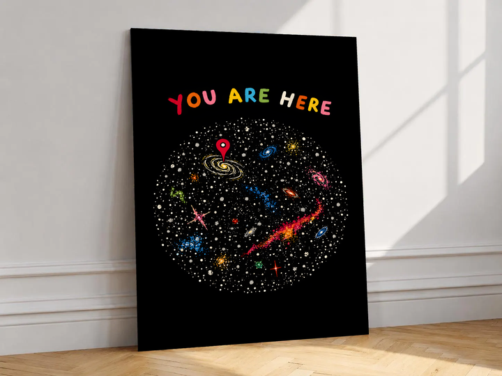 You Are Here multicolored lettering above a hand-drawn galaxy field with stars and a red location pin, shown unframed on a white wall above parquet flooring with a diagonal sun shadow