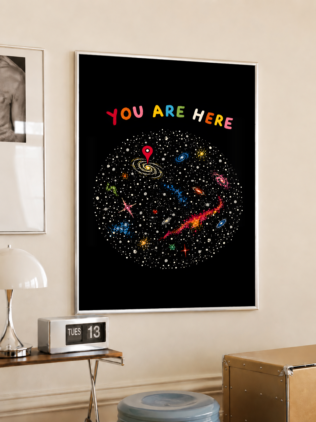 You Are Here multicolored lettering above a hand-drawn galaxy field with stars and a red location pin, styled in a light frame on a cream wall beside a white dome lamp and glass side table